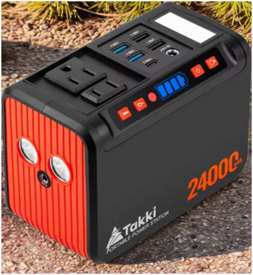 Black and red 2400C portable power station with AC and USB ports, shown on a rocky outdoor ground near pine needles, ready for off-grid use.