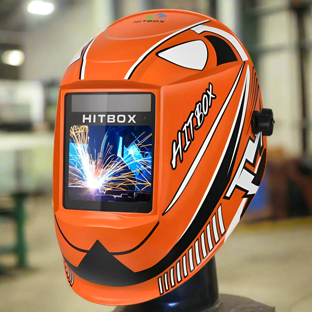 Orange HITBOX auto-darkening welding helmet with bold black-and-white accents, showcased inside a fab shop.